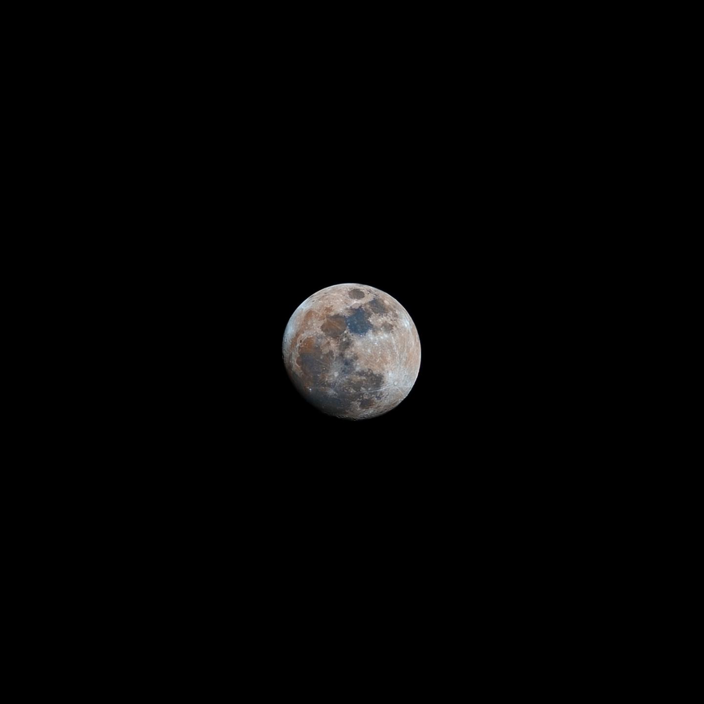 Photo of an almost full Moon from a slight distance, the bottom left limb is slightly in shadow. The mare, dark patches on the moon, stand out in different shades of grey, blue and rust